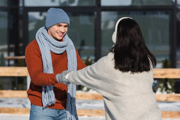 Feliz hombre en sombrero mirando borrosa novia mientras toma de la mano en pista de hielo - foto de stock