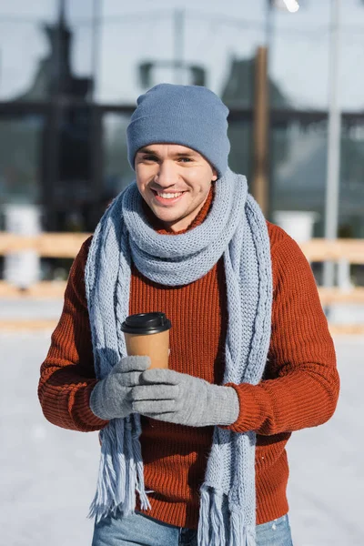 Happy young man in sweater, scarf and winter hat holding paper cup while skating on ice rink — Stock Photo