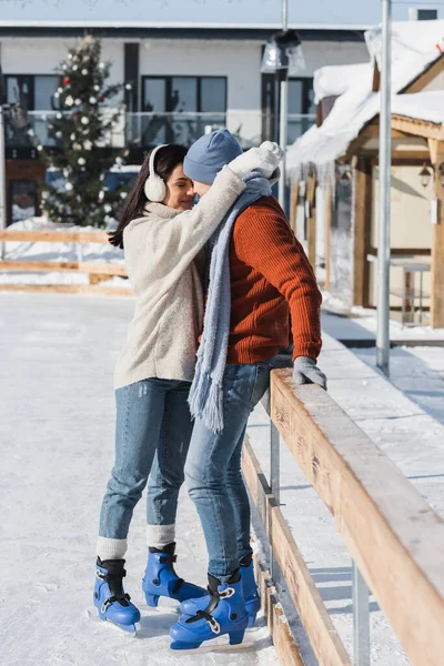 Pleine longueur de femme heureuse dans des cache-oreilles étreignant l'homme en chapeau d'hiver appuyé sur la bordure en bois sur la patinoire — Photo de stock