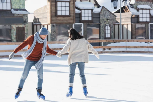 Longitud completa de la mujer joven en orejeras cogidas de la mano con el novio en pista de hielo - foto de stock