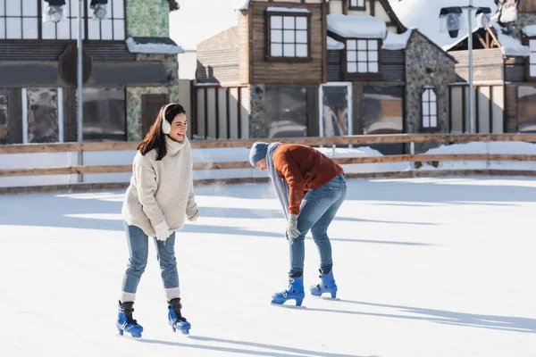 Pleine longueur de jeune femme dans les manchons d'oreille riant avec petit ami tout en patinant à l'extérieur — Photo de stock