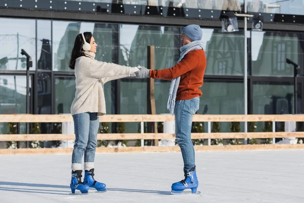 Full length of young woman in ear muffs holding hands with boyfriend on ice rink — Stock Photo