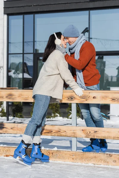 Full length of happy couple smiling while kissing near wooden border on ice rink — Stock Photo