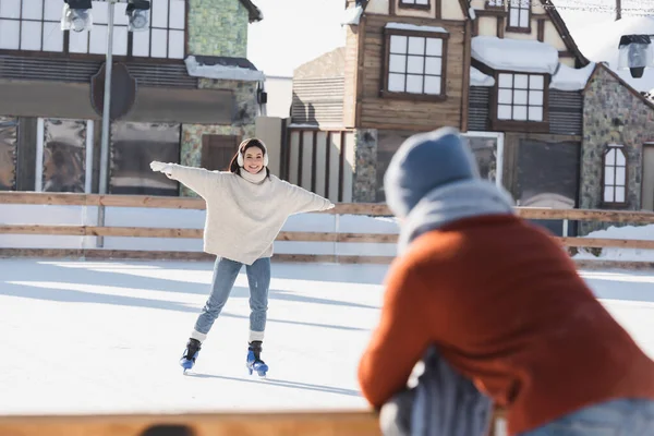 Lächelnde Frau, die auf der Eisbahn Schlittschuh läuft und ihren verschwommenen Freund im Vordergrund betrachtet — Stockfoto