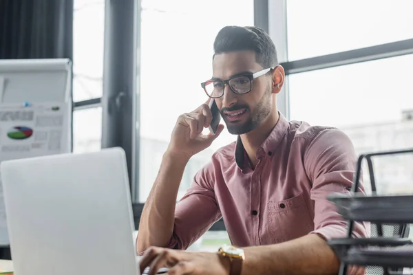 Homme d'affaires musulman positif parlant sur son téléphone portable et utilisant un ordinateur portable flou au bureau — Photo de stock