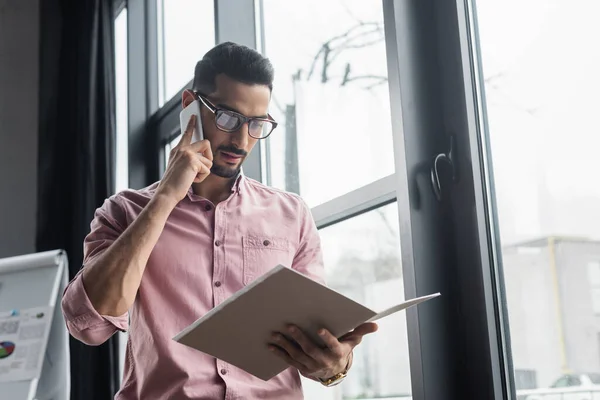 Muslimischer Geschäftsmann mit Brille spricht auf Smartphone und hält Papiermappe im Büro — Stockfoto