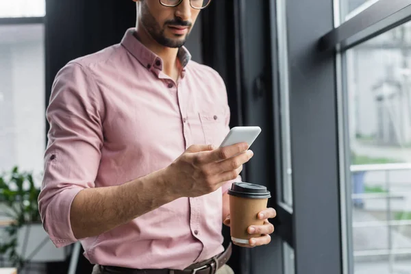 Vista recortada del hombre de negocios con taza de papel usando teléfono inteligente en la oficina - foto de stock