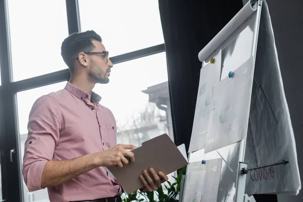 Side view of muslim businessman holding paper folder near flip chart in office — Stock Photo