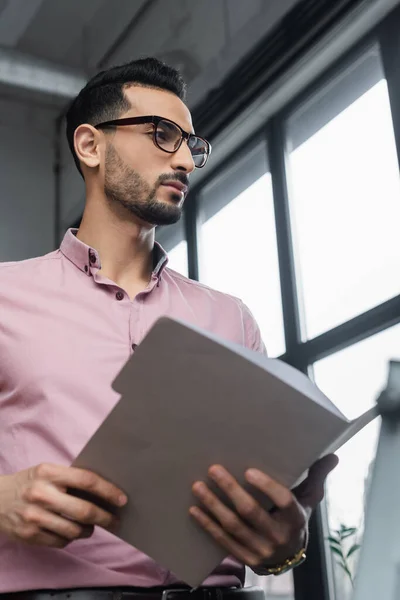 Low angle view of arabian businessman in eyeglasses holding blurred paper folder in office — Stock Photo