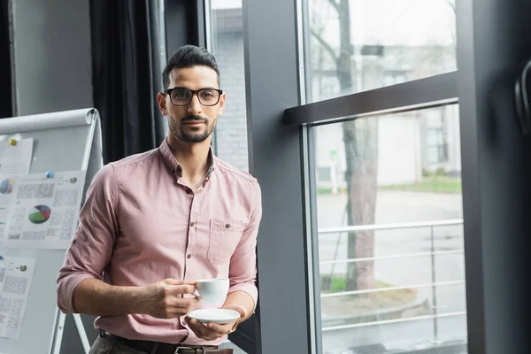 Joven hombre de negocios musulmán en gafas con taza y platillo en la oficina - foto de stock