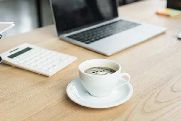 Tazza di caffè vicino computer portatile offuscata e calcolatrice in ufficio — Foto stock