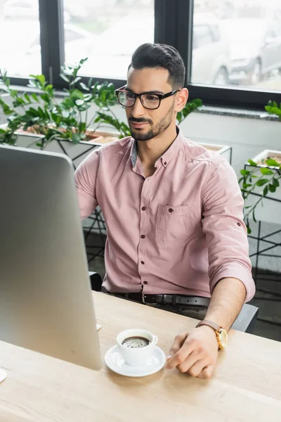 Uomo d'affari musulmano in occhiali da vista utilizzando il computer vicino a tazza di caffè in ufficio — Foto stock