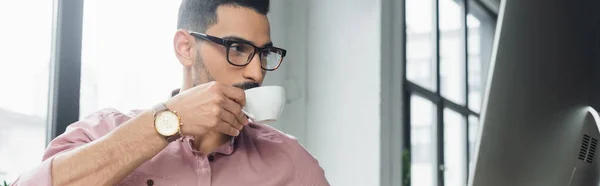 Молодой арабский бизнесмен пьет кофе и смотрит на монитор компьютера в офисе, баннер — стоковое фото