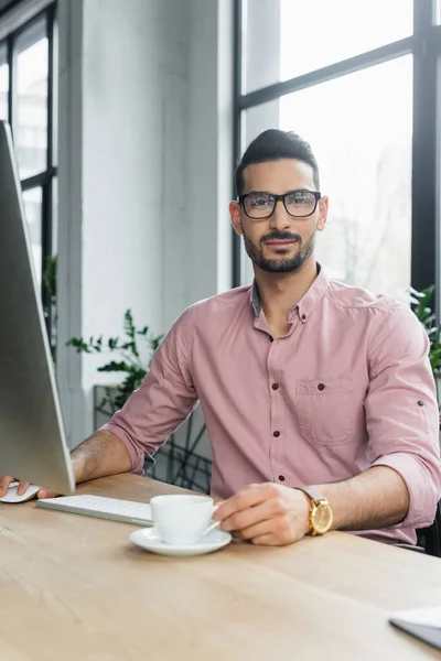 Hombre de negocios musulmán con taza mirando a la cámara cerca de la computadora en la oficina - foto de stock