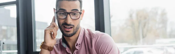 Молодой арабский бизнесмен в очках разговаривает по мобильному телефону в офисе, баннер — стоковое фото