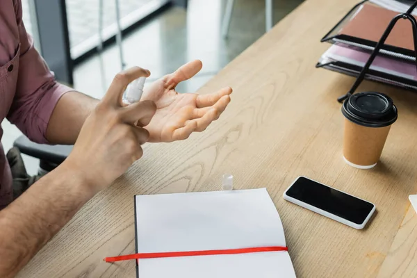 Ausgeschnittene Ansicht eines Geschäftsmannes, der Händedesinfektionsmittel in der Nähe von Smartphone, Coffee to go und Notebook im Büro versprüht — Stockfoto