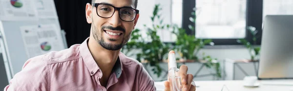 Fröhlicher muslimischer Geschäftsmann mit Brille und Händedesinfektionsmittel im Büro, Banner — Stockfoto