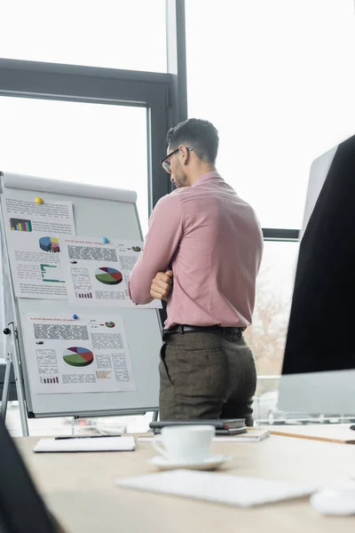 Arabischer Geschäftsmann mit Brille steht neben Flipchart mit Grafiken und Computer im Büro — Stockfoto