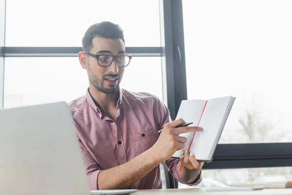 Empresário árabe em óculos apontando para notebook durante bate-papo por vídeo em laptop desfocado no escritório — Fotografia de Stock