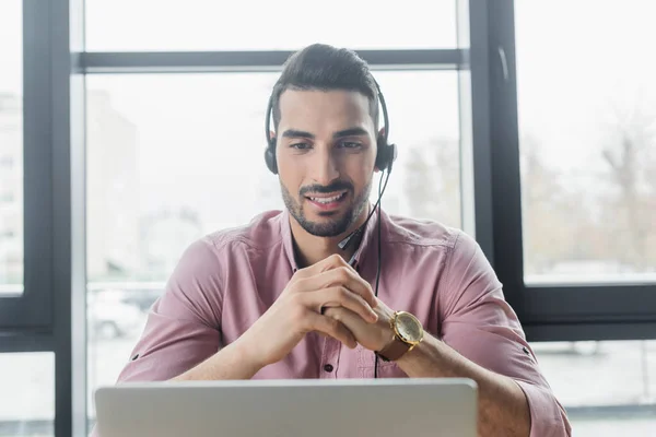 Sorridente uomo d'affari arabo in auricolare con videochiamata sul computer portatile offuscata in ufficio — Foto stock