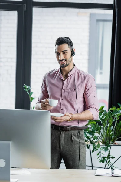 Homme d'affaires arabe positif dans un casque tenant tasse pendant l'appel vidéo sur l'ordinateur dans le bureau — Photo de stock