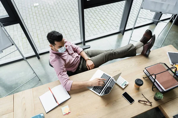 Vista aerea di uomo d'affari arabo in maschera medica utilizzando laptop vicino a caffè e notebook in ufficio — Foto stock