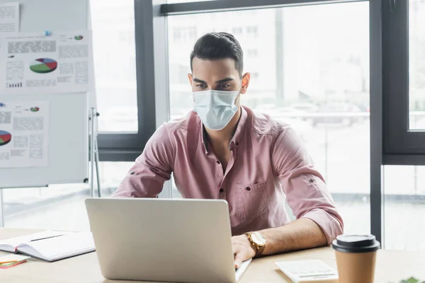 Arabian businessman in medical mask using laptop near coffee to go in office — Stock Photo