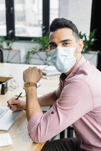 Arabian businessman in medical mask looking at camera near blurred laptop in office — Stock Photo