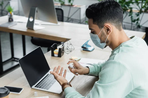 Arabian businessman in medical mask using hand sanitizer near devices in office — Stock Photo