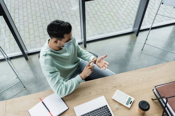 Arabischer Geschäftsmann in medizinischer Maske versprüht Händedesinfektionsmittel in der Nähe von Kaffee und Laptop im Büro — Stockfoto