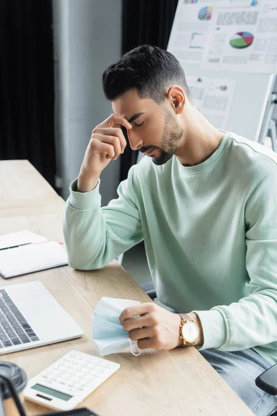 Uomo d'affari musulmano stanco che tiene maschera medica vicino al computer portatile in ufficio — Foto stock