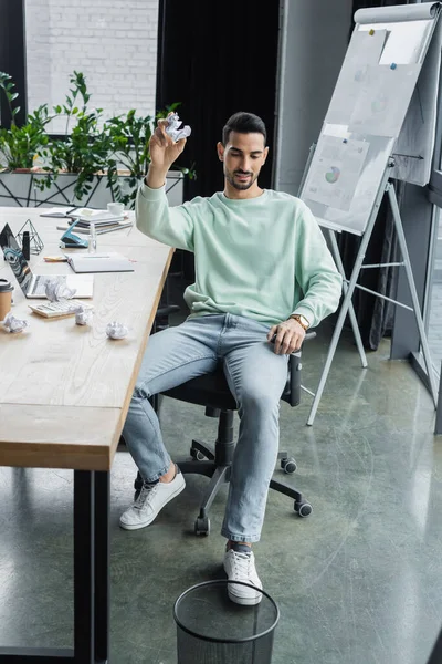 Joven empresario árabe sosteniendo papel arrugado cerca de mesa de trabajo y bote de basura en la oficina - foto de stock