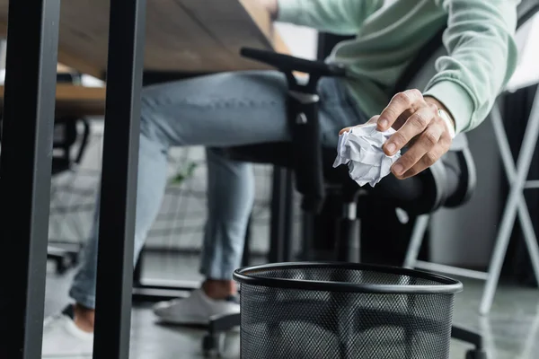 Cropped view of blurred businessman holding crumpled paper near trash can in office — Stock Photo