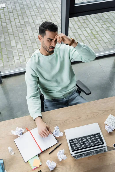 Visão superior do empresário árabe cansado olhando para laptop perto de notebook e papel amassado no escritório — Fotografia de Stock