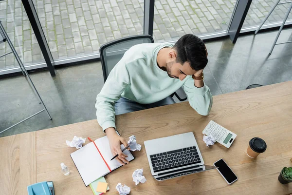 Vue aérienne d'un homme d'affaires musulman fatigué tenant un stylo près d'appareils, d'un ordinateur portable et d'un papier froissé au bureau — Photo de stock