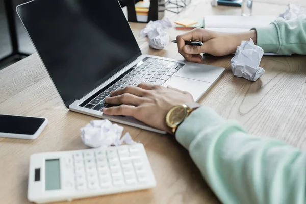 Ausgeschnittene Ansicht von Geschäftsmann mit Stift mit Laptop mit leerem Bildschirm in der Nähe zerknittertem Papier und verschwommenem Taschenrechner — Stockfoto