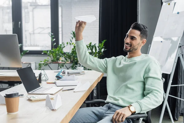 Lächelnder muslimischer Geschäftsmann mit Papierflieger in der Nähe von Geräten, Coffee to go und Taschenrechner auf dem Tisch — Stockfoto