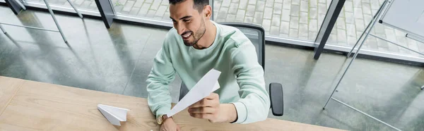 High angle view of smiling muslim businessman in casual clothes holding paper plane in office, banner — Stock Photo