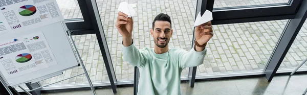 Vista ad alto angolo dell'uomo d'affari arabo con aerei di carta che sorridono alla fotocamera vicino al flip chart in ufficio, banner — Foto stock