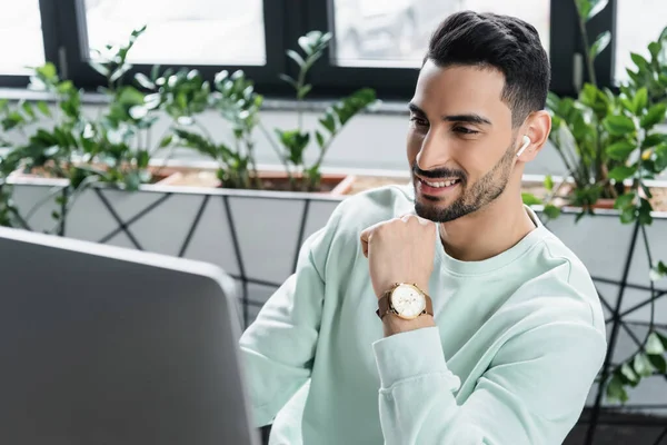 Homme d'affaires arabe positif dans un écouteur sans fil en utilisant un ordinateur flou — Photo de stock