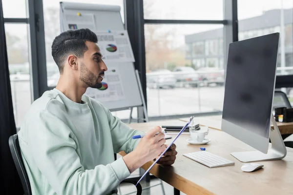 Side view of muslim businessman writing on clipboard near computer with blank screen in office — Stock Photo