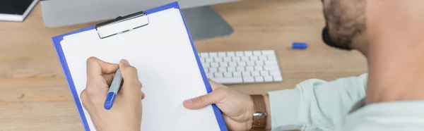 Cropped view of businessman writing on clipboard near blurred computer keyboard in office, banner — Stock Photo