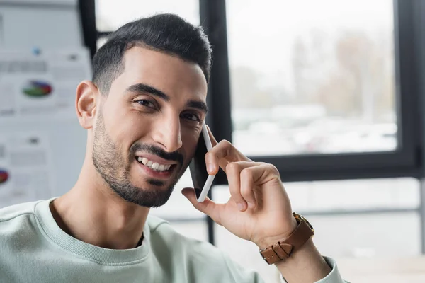 Homem de negócios muçulmano sorridente falando no smartphone e olhando para a câmera no escritório — Fotografia de Stock