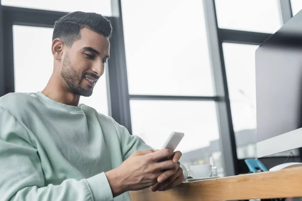 Young arabian businessman using smartphone near computer monitor in office — Stock Photo