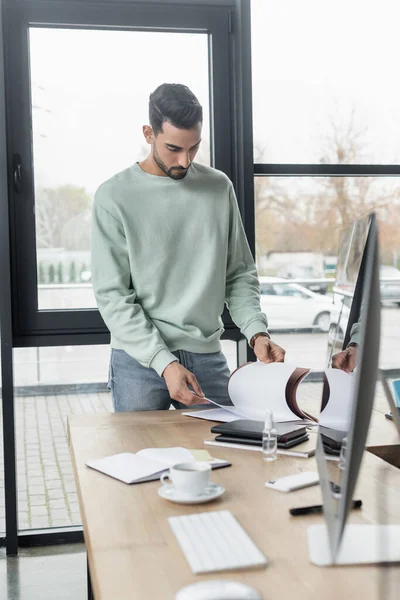 Muslimischer Geschäftsmann schaut sich im Büro Papiere in der Nähe von Händedesinfektionsmittel und Computer an — Stockfoto