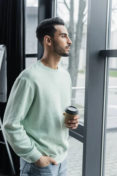 Vista lateral del joven empresario árabe sosteniendo café para acercarse a la ventana en la oficina - foto de stock