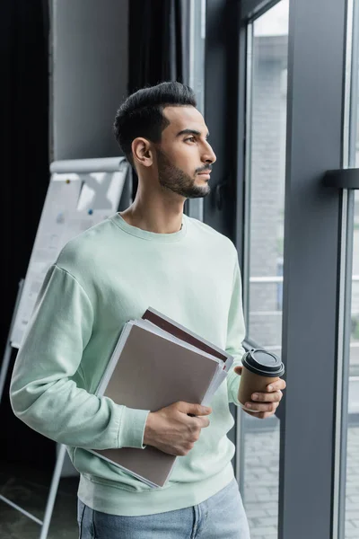 Young muslim manager in casual clothes holding paper folders and paper cup near window — Stock Photo