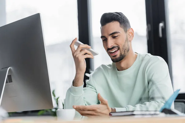 Uomo d'affari musulmano sorridente che registra il messaggio vocale sullo smartphone vicino al computer e al caffè in ufficio — Foto stock