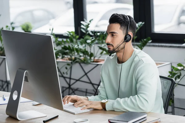 Joven hombre de negocios árabe en auriculares con computadora cerca de la taza y cuadernos en la oficina - foto de stock