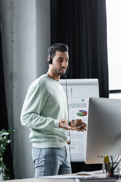 Smiling muslim businessman in headset having video call on computer near blurred flipchart — Stock Photo